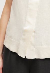 Krämfärgad stickad topp med ribbad nederkant och sid-slitsar. Tyget har en slät textur, med en minimalistisk design och inga synliga mönster.