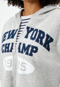 Grau zip-up Hoodie mit navyblauer Stickerei "NEW YORK CHAMP" und der Zahl "1985", kombiniert mit einem gestreiften Hemd darunter.