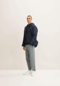 Chaqueta acolchada azul marino sobre una sudadera azul oscura, combinada con jeans gris claro y zapatillas blancas. Texturas suaves y diseño casual.