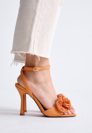 Pie con sandalia naranja de tacón alto con correa en el tobillo y gran flor de tela, combinado con pantalones blancos cortados y deshilachados.