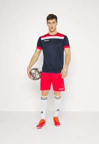 Marineblaues und rotes Sporttrikot mit weißen Akzenten, kombiniert mit roten Shorts. Das Modell hält einen gemusterten Fußball, trägt weiße Socken und rote Fußballschuhe.