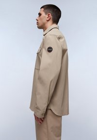 Camisa de manga larga beige que presenta un cuello con botones, un bolsillo en el pecho y un parche redondo en la manga. Tejido suave con un corte relajado.