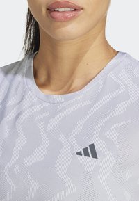 Camiseta deportiva blanca con un patrón texturizado en zigzag, cuello redondo y un pequeño logo gris en el lado izquierdo, fabricada con material sintético.