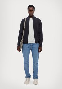 Marineblaues Zip-up-Jackett, weißes T-Shirt, blaue Jeans und helle Sneaker. Das Model hält eine graue Umhängetasche. Schlichtes, lässiges Outfit mit lockerem Sitz.