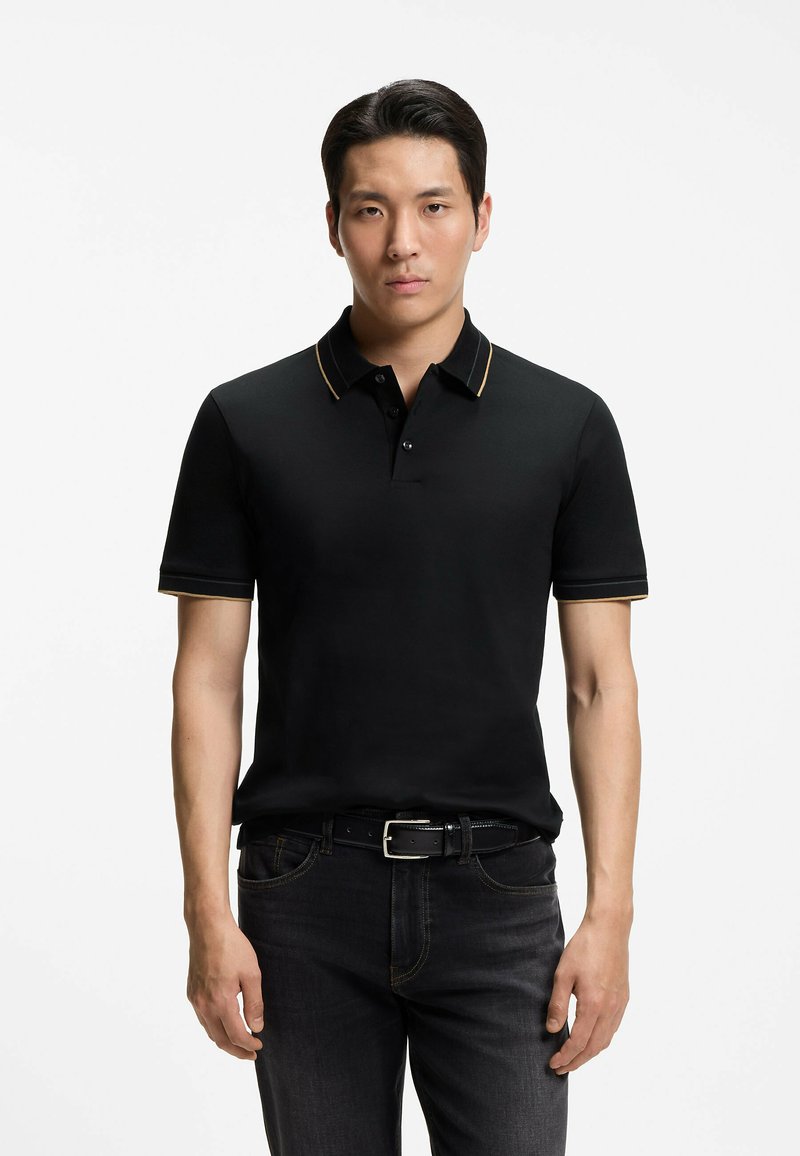 Jeune homme portant un polo noir à manches courtes avec une subtile bordure dorée, un jean foncé et une ceinture noire, debout sur un fond blanc.