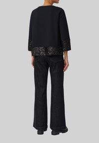 Schwarzes Oberteil mit glitzerndem Pailletten-Saum und dreiviertelÄrmeln, kombiniert mit dunklen, weit geschnittenen Jeans und schwarzen Stiefeln. Insgesamt glatte Textur.