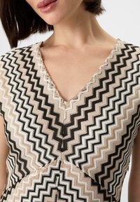 Strickoberteil mit V-Ausschnitt und Zickzackmuster in Beige, Weiß und Schwarz. Strukturiertes Design mit kontrastierenden Farben und sichtbaren Nähten.