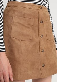 Bruine suède mini-rok met twee voorzakken en knoopsluiting. Heeft een gladde textuur en een recht afgesneden ontwerp. Verticale stikseldetails.