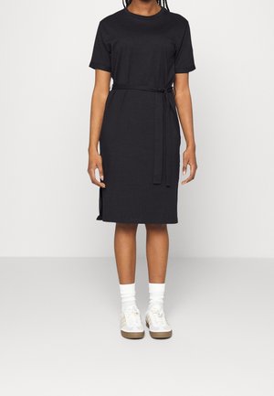 Kvinde iført en knælang sort kjole med korte ærmer og bånd i taljen, hvide strømper og hvide sneakers, stående mod en ensfarvet baggrund.