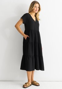 Black, knee-length dress featuring a V-neck, short sleeves, tiered design, and side pockets; worn with brown, two-strap sandals.