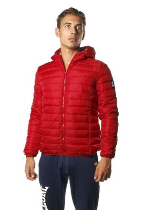 Homme posant avec une veste matelassée rouge à capuche et un pantalon de jogging bleu marine sur un fond blanc uni.