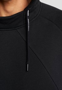Sudadera negra con un cuello alto, que presenta una tela texturizada, sutiles detalles de costura y un cordón con marca en la punta.