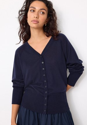 Vrouw met krullend haar die gouden oorringen draagt, een marineblauwe gebuttonde cardigan en een bijpassende geplooide rok, staand tegen een witte achtergrond.