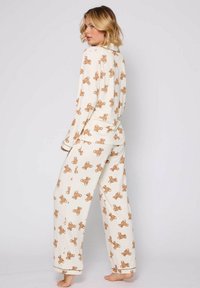 White pajama set featuring repeated light brown teddy bear print. Long-sleeved top with collar, and loose-fitting pants with cuffed hems.