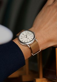 Käekell valge dialiga, mustade numbrite ja beeži nahkriiuliga, kantud vasakul randmel, näitab aega 8:11 koos kuupäeva suurendajaga kell 3.