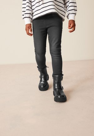 Sorte læder ankelstøvler med en kraftig sål, spænde detalje og lynlåse. Båret med mørke jeans og en hvidstribet top. Glat tekstur.
