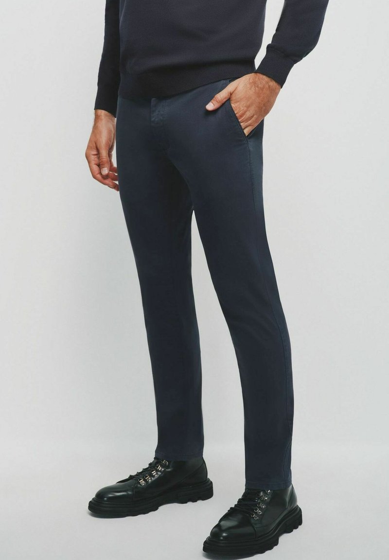 Granatowe, wąskie spodnie uszyte z gładkiego materiału, z kieszeniami z przodu, noszone z czarnymi sznurowanymi botkami na masywnych podeszwach.