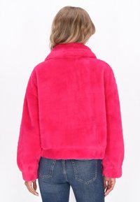 Kvinna med vågig blont hår klädd i en ljusrosa fuskpälsjacka och blå jeans, stående vänd bort från kameran mot en vit bakgrund.