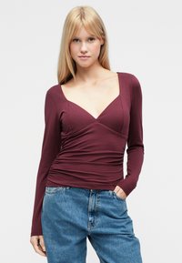 Burgundy long-sleeve top featuring a deep V-neck, ruched detailing at the waist, and smooth fabric, paired with high-waisted denim jeans.