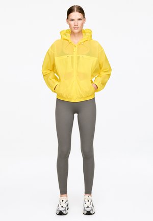 Kvinde stående med front mod kamera iført gul hættejakke, grå leggings og hvide sportssko med hænderne i jakkelommerne.