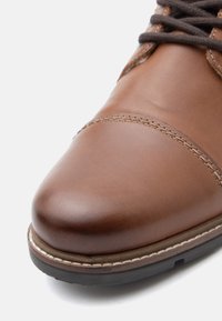 Brązowy skórzany but z zaokrąglonym czubkiem, gładką fakturą, kontrastowym przeszyciem wzdłuż krawędzi oraz gumową podeszwą dla lepszej przyczepności.