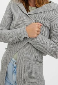 Hrubě pletený kardigan světle šedé barvy s velkou kapucí, předními kapsami a žebrovanými manžetami. Texturovaný, teplý materiál s volným střihem.