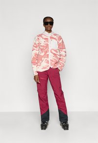 Fleecejakke med pink og hvid swirl-mønster, lynlåslomme; kombineret med vinrøde skibukser med navy-detaljer og lynlåse. Sorte skistøvler.