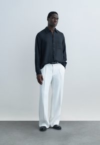 Mladý muž stojí pred neutrálnym pozadím, oblečený v voľnej čiernej košeli na gombíky, bielych plisovaných nohaviciach a čiernych topánkach, ruky v vreckách.