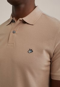Beige polo skjorta i texturerad tyg, med en knapp-ned krage och en liten broderad fågel-logotyp på bröstet.