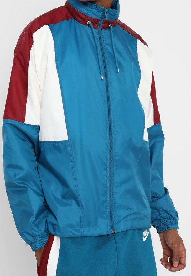 Veste coupe-vent teal avec des accents blancs et bordeaux, fermeture éclair intégrale sur le devant, col haut et poignets élastiques ; matériau lisse et léger.