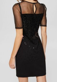 Černé šaty s průhlednými krátkými rukávy, zdobené flitry na živůtku, které se směrem k lemu ztrácejí, s designem výstřihu ve tvaru klíčové dírky na zádech.