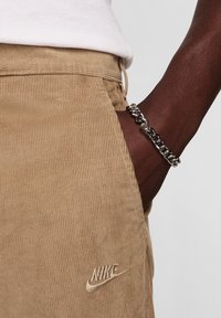Pantalones de pana marrón con rayas verticales y un logo de Nike. Una pulsera de cadena plateada envuelve una muñeca, con eslabones entrelazados.