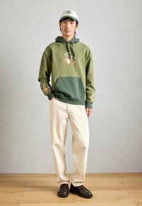 Νεαρός άνδρας με πράσινη κουκούλα με γραφικά σχέδια, κρέμα παντελόνια, σκούρα καφέ παπούτσια και λευκό καπέλο μπέιζμπολ, που στέκεται σε ξύλινο δάπεδο μπροστά σε γκρίζο τοίχο.