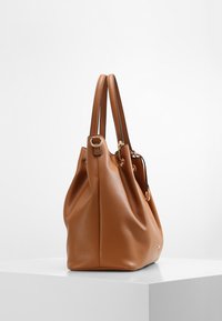 Braune strukturierte Lederhandtasche mit zwei Griffen, goldfarbener Hardware und einem lässigen Design, auf einem weißen Podest platziert.