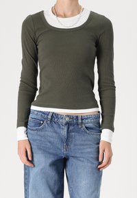 Maglietta verde oliva a maniche lunghe con scollatura rotonda e texture a coste, indossata sopra una camicia bianca. Abbinata a jeans baggy azzurri.