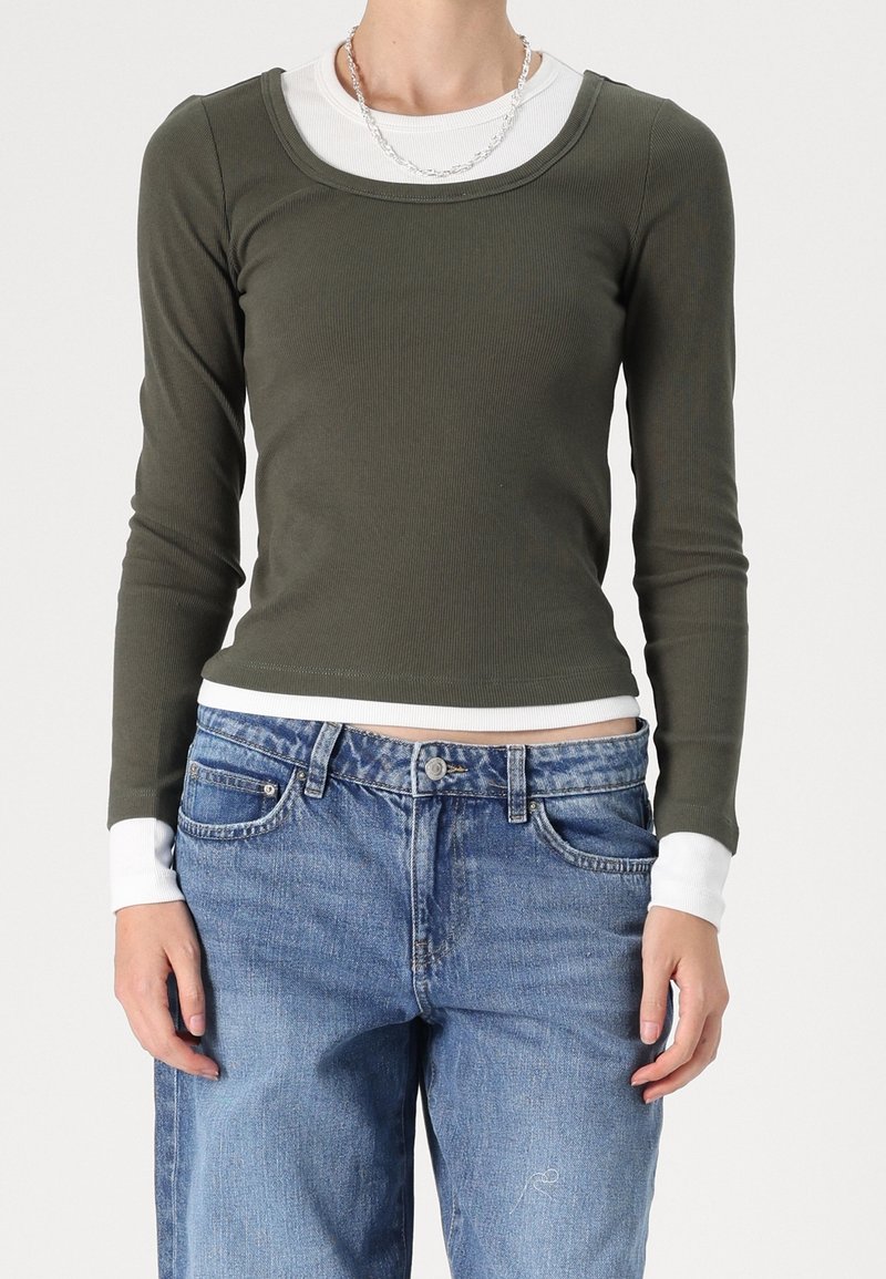 Maglietta verde oliva a maniche lunghe con scollatura rotonda e texture a coste, indossata sopra una camicia bianca. Abbinata a jeans baggy azzurri.