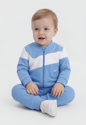 Småbarn med lyst hår, der sidder med korslagte ben iført en blå og hvid jakke med lynlås og hvide sneakers, smilende let til siden.