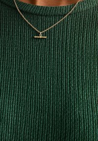 Goldene Halskette mit einem horizontalen Balkenanhänger, der auf einem strukturierten, dunkelgrünen, gerippten Stoff liegt. Die Kette ist zart und dünn.