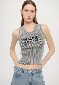 Ung kvinne med rett hår som har på seg en ermeløs grå og svart stripete topp med "MOSCHINO JEANS" tekst og lyseblå jeans.