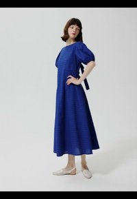 Žena stoji s rukom na bokovima, nosi plavu midi haljinu s rukavima na napuhavanje i bijele slingback cipele s niskom petom, na jednobojnoj pozadini.