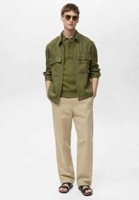 Olive green jacket with two chest pockets over a ribbed green polo, paired with beige trousers and black sandals. Smooth textures displayed.