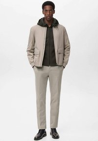 Jachetă ușoară bej peste un hanorac verde închis cu fermoar, combinată cu o pereche de pantaloni bej deschis și pantofi negri. Design minimalist, textură netedă.