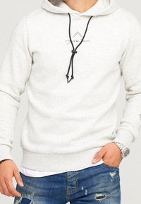 Mann trägt einen hellgrauen Kapuzenpullover mit schwarzen Kordeln, ein weißes Unterhemd, ausgebleichte blaue Jeans und eine silberne Armbanduhr, Hand in der Tasche.