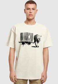 Cremefarvet oversized t-shirt med et sort stregkode-design med dråber og en silhuet af en kat på fronten. Korte ærmer.