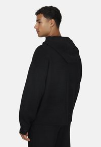 Mężczyzna w luźnym czarnym swetrze z dzianiny i dopasowanych spodniach, pokazany od tyłu i lekko obrócony w lewo na jednolitym tle.