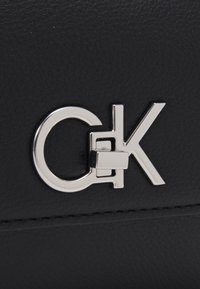 Zwart gestructureerd leren product met een zilveren sluiting met een cirkelvormig ontwerp, waarin de letters "C" en "K" in een modern lettertype zijn verwerkt.