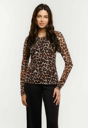 Mladá žena s dlouhými tmavými vlasy, oblečená v dlouhém rukávu s hnědočerným leopardím vzorem a černých kalhotách, stojící před bílým pozadím.