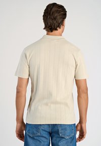 Beige polotröja med korta ärmar, som har ett strukturerat geometriskt mönster. Kombinerad med blå denim-jeans. Detaljerad sömnad synlig.