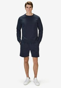 Marinblå sweatshirt med rund hals och långa ärmar; matchas med marinblå shorts, båda i ett slätt material. Vita sneakers fulländar outfiten.