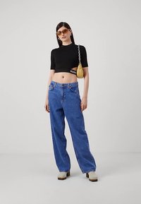 Melna apgriezta t-krekla ar logotipu, zilas platām kājām džinsi, bēša platformas klampsandales un oranžas saulesbrilles. Dzeltena rokassoma ar ķēdes siksnu.